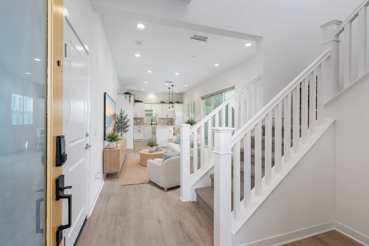 Your Entry from the Front Door. Guest Bath to the right. View the Open Living Space: Living Room, Dining Area, Kitchen. Door to Garage on the left, across from the stairs. Door to the Main Floor Primary Suite is just past the TV on the left. Photo is virtually staged.